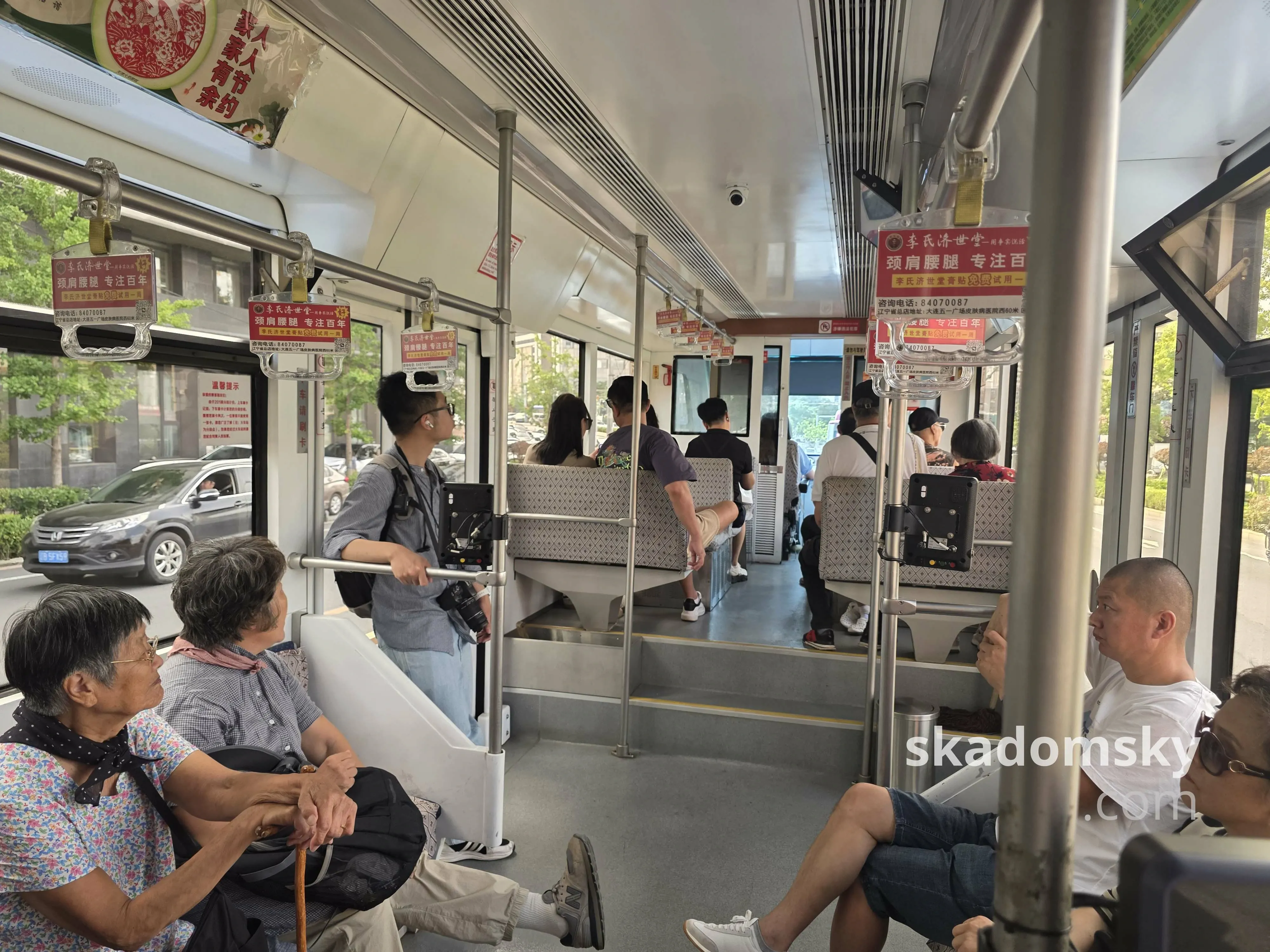 有轨电车车头车厢内部。不少老人坐在两侧的座位上，左侧车门处一个挂着相机的男人靠着栏杆。